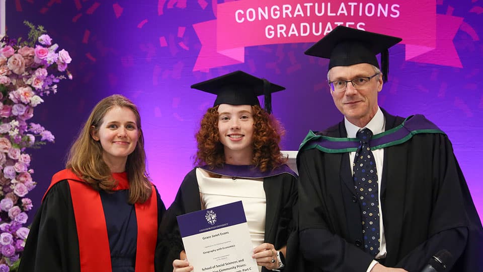 Grace holding her award in her graduation cap and gown in front of a sign that says Congratulations Graduate in Loughborough colours.